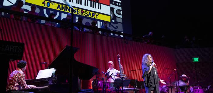 Black woman in black dress singing in front of a band on stage with a huge colorful banner behind them.
