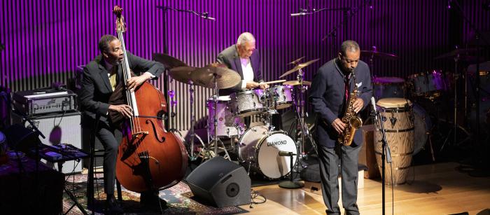 Three elderly Black men in suits playing drums, bass, and sax on stage with purple background. 