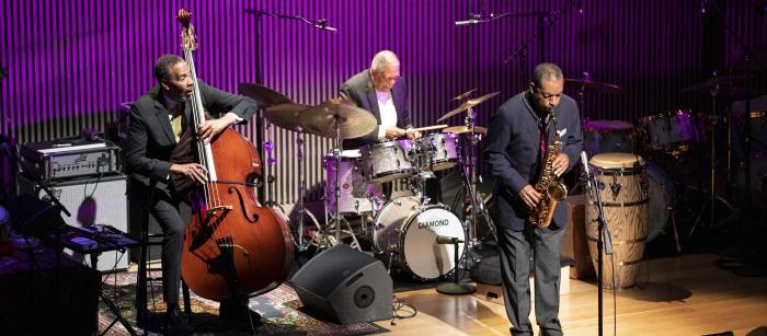 Three elderly Black men in suits playing drums, bass, and sax on stage with purple background.