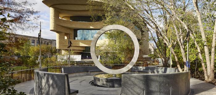 a view of the National Native American Veterans Memorial, which is a large circle on its side on top of a drum shape. The exterior of the National Museum of the American Indian is visible in the background