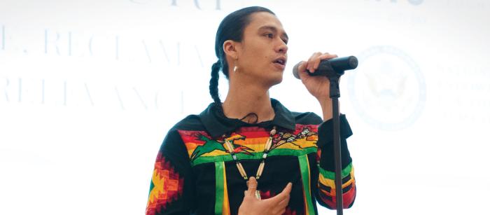 Native man with hair in braid, wearing colorful traditional clothes, at a microphone on stage.