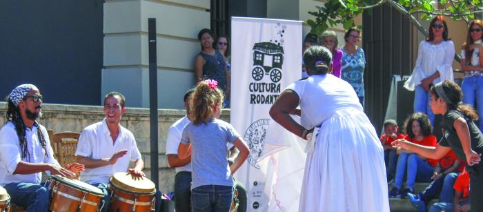 Woman in white dress teaching children to dance outdoors, with men playing drums in background.