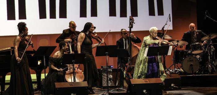 Group of women playing stringed instruments on stage, the one on the left wearing a gold jacket.wearing a