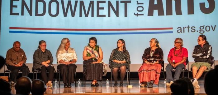 A group of people are sitting on stage, woman in the middle holding a microphone, screen showing National Endowment for the Arts behind them. 