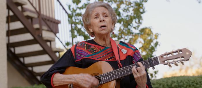 Woman with gray hair playing a guitar outside in front of a staircase. 