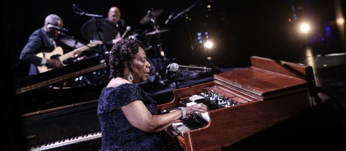 Older Black woman seated and playing the organ on stage.