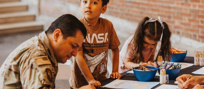 A man in military uniform and two children sit at a table coloring with crayons.