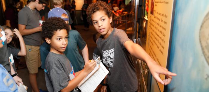 Two boys engage in an art exhibit at a museum. In the foreground, one child points at an information board while another takes notes.