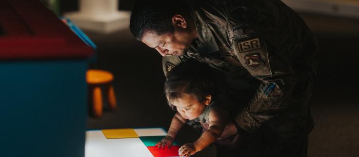 A man dressed in military uniform holds a child to allow access to a brightly lit table featuring colored squares.