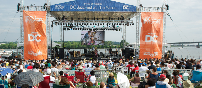 An audience sits and watches musicians performing on an outdoor stage in summer. 