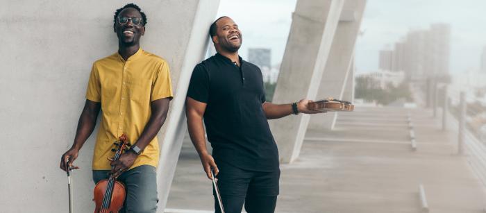 Two men stand against a pillar and laugh while holding violins