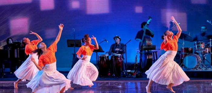 Four dancers in orange tops and white skirts perform. There is also a band playing upstage.