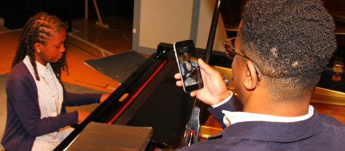 A man uses his cell phone to record a young woman playing the piano.
