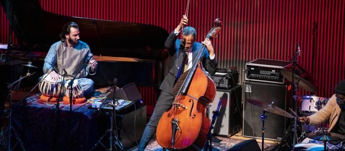 Three men onstage playing music, including a drummer, a bassist, and a man playing table, which are a type of hand drum.