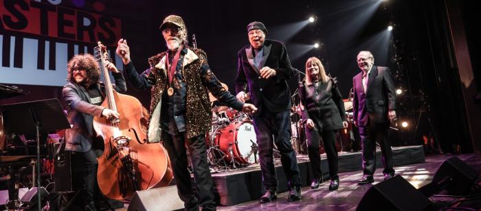 Three men and a woman smile and walk single-file onstage while musicians play behind them.