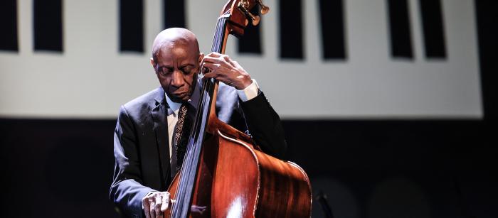 A man plays upright bass onstage in front of a backgroup of an illustration of piano keys.