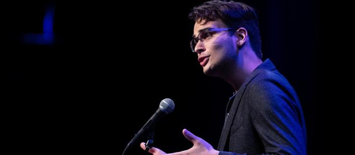 A young man speaks into a microphone on a darkened stage.