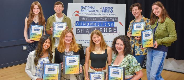 A group of teenagers smile and pose in front of a sign for the Musical Theater Songwriting Challenge. They each hold a binder with their name and the name of their winning song.