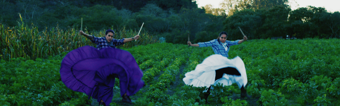 Two woman in a field on a farm dancing with sticks, wearing white and blue frilly dresses and plaid shirts.