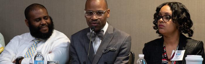 Two African American men and an African American women on a panel - the men in the center is speaking into a mincrophone