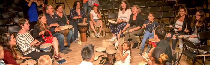 Large indoor drum circle of men and women with a small child in the middle on the floor m