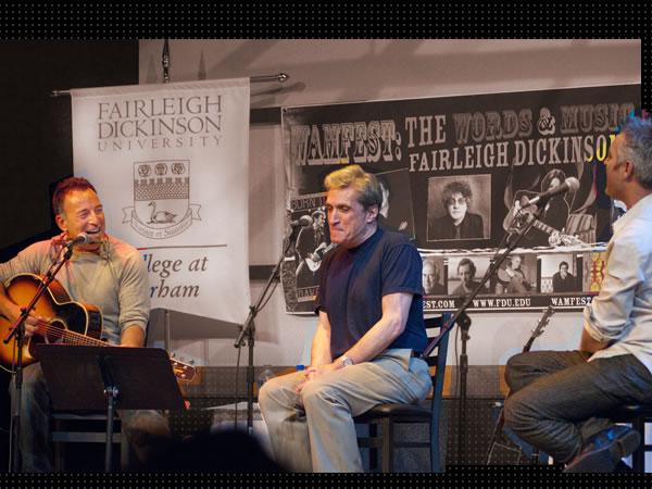Musician Bruce Springsteen laughs as he reacts to poet Robert Pinsky and author Wesley Stace on stage at the Words and Music Festival, 2010.  The backdrop is a WAMFEST poster with images of participating artists.