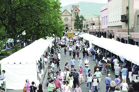 Crowds of people walking down the street and looking into various white tents lined up along the curb; an old church in the background.