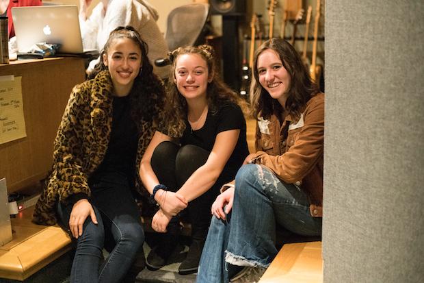 Three young women sit close to one another in a recording studio, next to a desk, smiling for a photo.