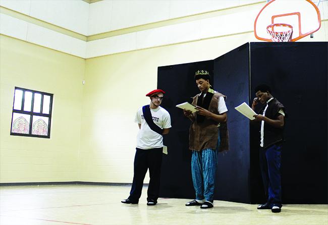 Shakespeare in American Communities Three young men wearing costumes read from scripts in a gymnasium