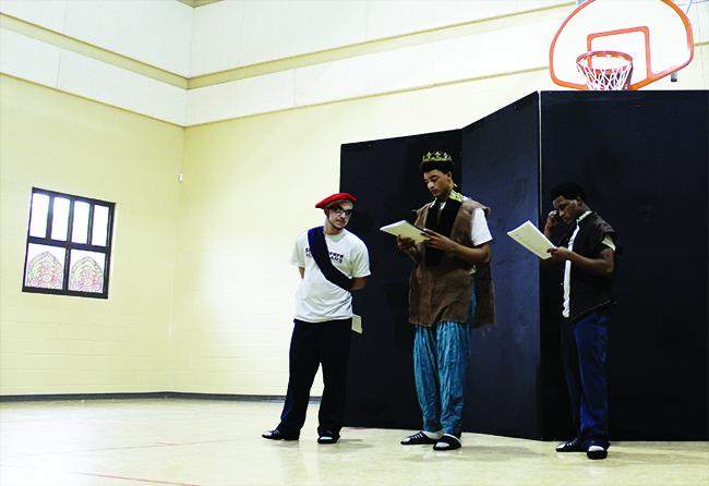 Three young men wearing costumes read from scripts in a gymnasium