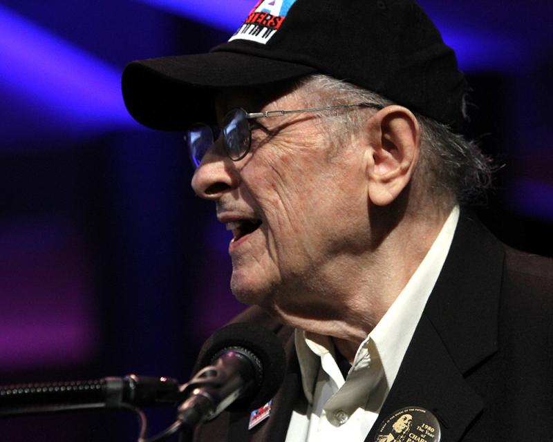 A man in glasses and a baseball hat speaks from a podium