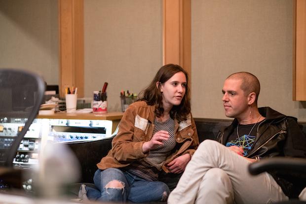 A young woman sits next to a bald man on a couch in a recording studio, speaking to him.