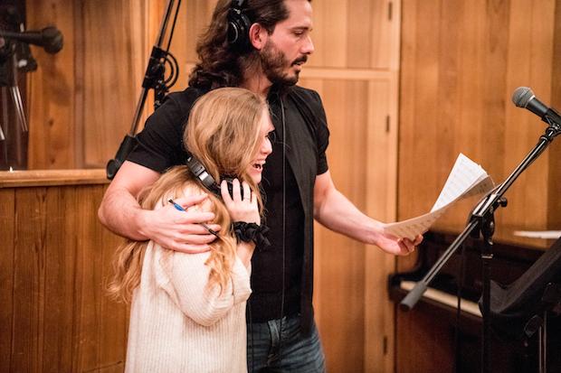 A man with a beard with his arm around a laughing young woman, both singing in front of a microphone in a recording studio.