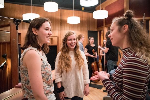 Three young women, talking to each other in a recording studio, smiling. There are four musicians in the background, two women and two men, holding their instruments.