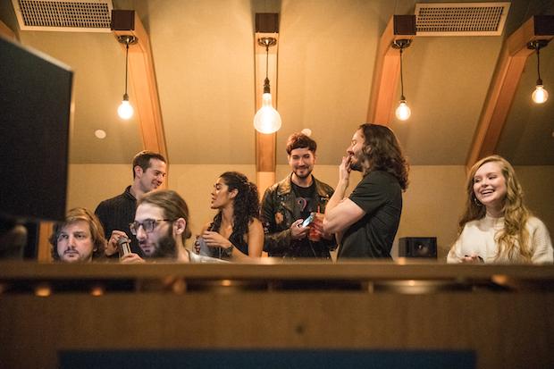 A group of five men and two women stand behind a desk in a recording studio, laughing and working.