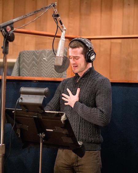 A man in a gray sweater stands behind a suspended mic in a recording studio, singing from sheet music on a music stand.