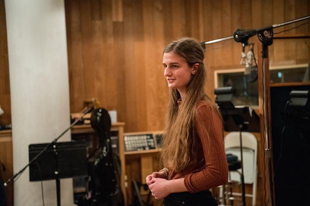 A young woman stand center frame in a recording studio full of recording equipment.