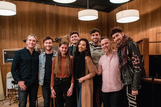 A group of six men and two women pose for a photo in a recording studio, arms around each other, smiling.