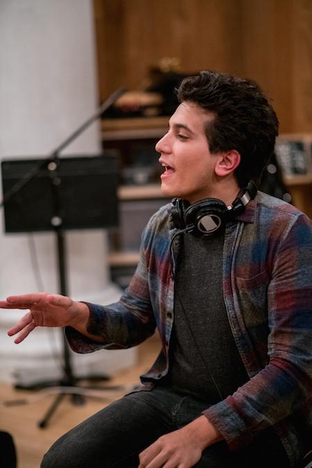 A young man in a flannel with headphones around his neck, speaking in a recording studio.