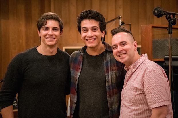 A man singing in the foreground of a recording studio. Two young men sit, blurry, in the background.