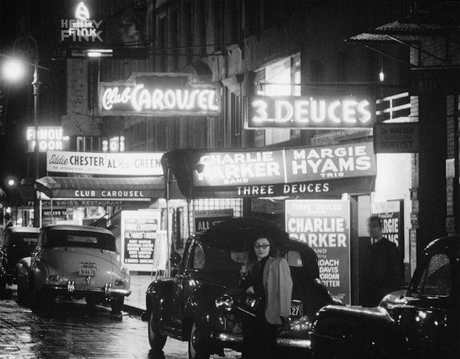 a view of a row of nightclubs on 52nd street with the names of each club lit up in neon and other lights. You can see some people and a row of cars, and some of the evening bills at each club