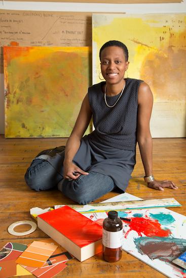 an african american woman with very short hair sits on a wooden floor in front of painted canvases with paint chips and other paraphenalia in front of her