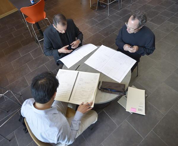 A view from on high of a trio of men sitting around a table looking a music scores