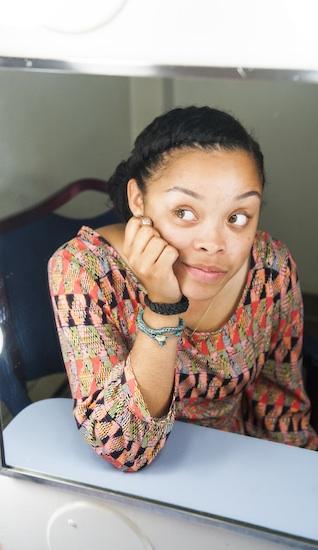 A young black woman looks sideways and is reflected in a dressing room mirror