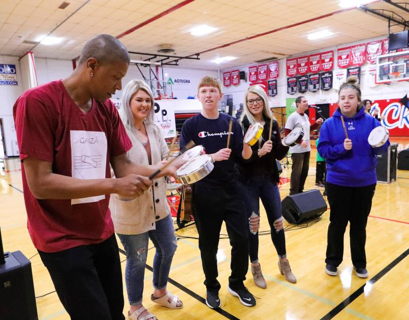 Samba Sam A man shows a group of teenagers in a high school gym how to play a Brazilian instrument