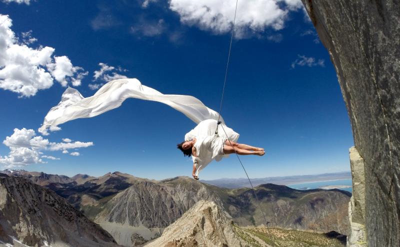 A dancer with flying white skirt floats above the Sierra Nevada Mountains while tethered to the mountain