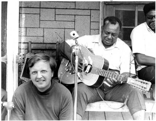 A man sits smiling on a porch step while a man plays guitar into a microphone while sitting behind him