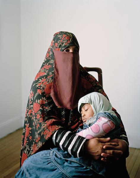 a woman in a floral print burqa sits in a chair cradling her young daughter who wears play clothes and a headscarf