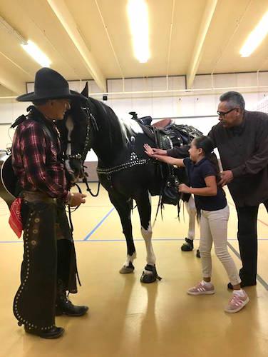 a man in Western wear holds the reins of a horse while a young girl pats the side of the horse