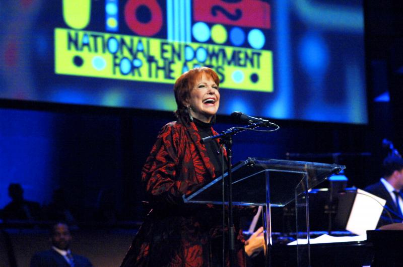2010 NEA Jazz Master Annie Ross A woman with short red hair and a bright smile stands at a podium and microphone with a colorful sign in the background.